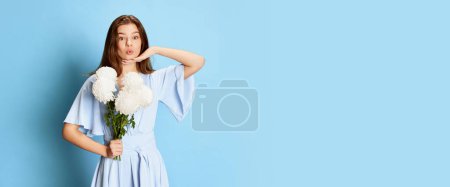 Téléchargez les photos : Portrait de jeune fille en jolie robe tendre posant avec des fleurs blanches sur fond bleu. Flyer. Concept de fête, fête, journée des femmes, émotions, vacances, bonheur. Publicité - en image libre de droit