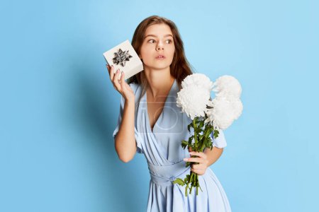 Téléchargez les photos : Portrait de jeune fille en robe tendre mignon posant avec des fleurs blanches et boîte cadeau sur fond bleu. Le 8 mars. Concept de fête, fête, journée des femmes, émotions, vacances, bonheur. Publicité - en image libre de droit