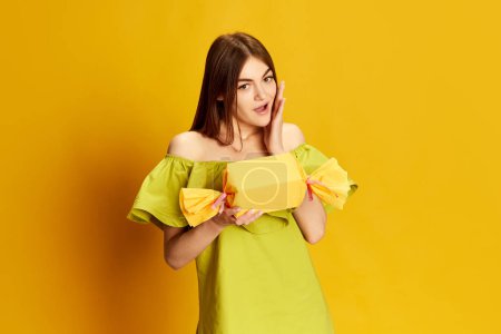 Photo for Surprise. Portrait of young beautiful girl in cute dress posing with present box over yellow background. Concept of celebration, party, womens day, emotions, holiday, birthday, happiness. Ad - Royalty Free Image