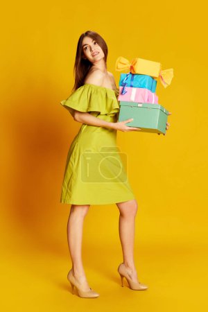 Photo for Portrait of young beautiful girl in cute dress posing with many present boxes over yellow background. Concept of celebration, party, womens day, emotions, holiday, birthday, happiness. Ad - Royalty Free Image