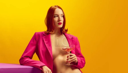 Photo for Drink aesthetics. Young redhead woman in pink jacket holding glass with champagne over yellow background. Concept of holiday, party, drink. Complementary colors. Copy space for ad. Pop art - Royalty Free Image