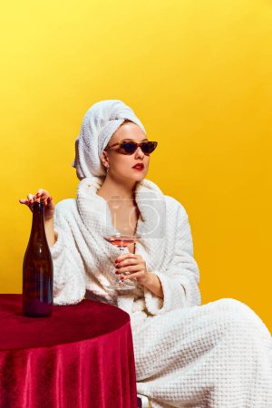 Foto de Mujer joven en albornoz sentada y bebiendo champán sobre fondo amarillo. Fiesta en casa. Relajación. Concepto de fiesta, fiesta, bebida. Colores complementarios. Copia espacio para el anuncio. Arte pop - Imagen libre de derechos
