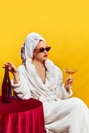 Téléchargez les photos : Jeune femme en peignoir assise et buvant du champagne sur fond jaune. Je me sens détendu. Concept de vacances, fête, boisson. Couleurs complémentaires. Espace de copie pour l'annonce. Pop art - en image libre de droit