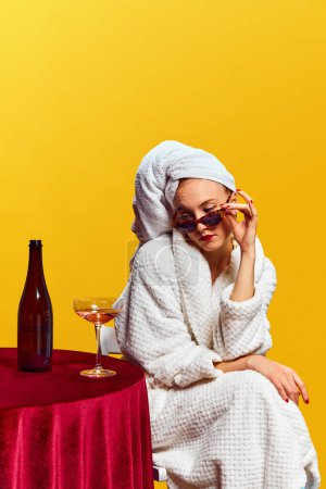 Foto de Mujer joven en albornoz sentada y bebiendo champán sobre fondo amarillo. Celebración. Concepto de fiesta, fiesta, bebida. Colores complementarios. Copia espacio para el anuncio. Arte pop - Imagen libre de derechos