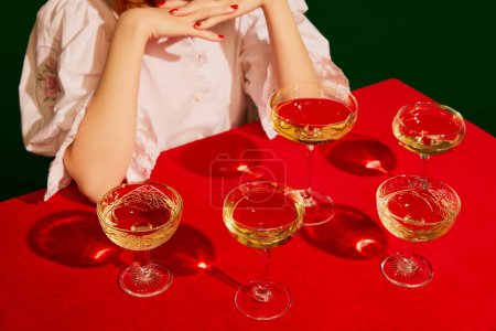 Téléchargez les photos : Image recadrée d'une femme en robe blanche assise à la table avec des verres à champagne sur une nappe rouge sur fond vert. Concept de fête, boisson. Couleurs complémentaires. Espace de copie pour l'annonce. Pop art - en image libre de droit