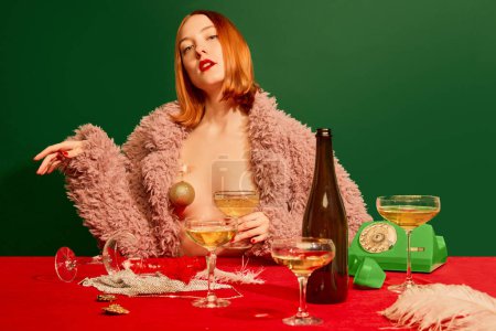 Téléchargez les photos : Fille rousse en manteau de fourrure rose assis à la table avec du champagne sur fond vert. Fête du Nouvel An. Concept de vacances, fête, boisson. Couleurs complémentaires. Espace de copie pour l'annonce. Pop art - en image libre de droit