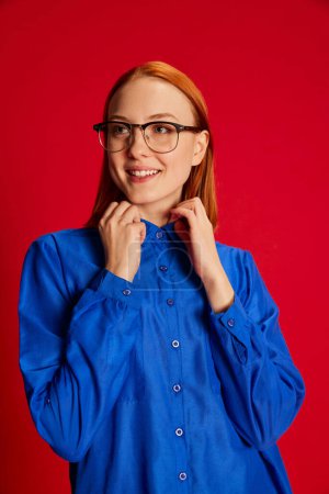 Foto de Retrato de una joven pelirroja sonriente con camisa azul y gafas posando sobre fondo rojo. Me siento bien. Concepto de juventud, belleza, moda, estilo de vida, emociones, expresión facial. Anuncio - Imagen libre de derechos