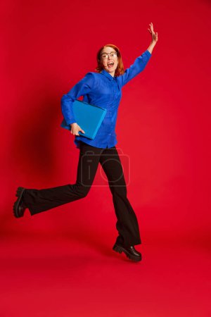 Téléchargez les photos : Jeune fille rousse en chemise bleue et lunettes posant dans un saut avec ordinateur portable sur fond rouge. Étudiant réussi. Concept de jeunesse, beauté, mode, mode de vie, émotions, expression faciale. Publicité - en image libre de droit