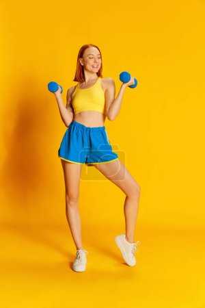 Photo for Portrait of young redhead girl in sportswear training with sports equipment over yellow background. Hands exercises. Concept of youth, beauty, sport, lifestyle, emotions, facial expression. Ad - Royalty Free Image