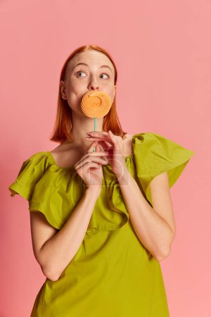 Photo for Portrait of young beautiful girl in cute dress posing with candy over pink background. Good mood and positive energy. Concept of youth, beauty, fashion, lifestyle, emotions, facial expression. Ad - Royalty Free Image
