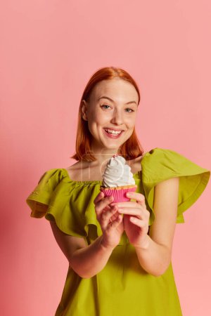 Téléchargez les photos : Portrait de jeune belle fille en robe mignonne posant avec cupcake sur fond rose. Doux Jésus. Concept de jeunesse, beauté, mode, mode de vie, émotions, expression faciale. Publicité - en image libre de droit