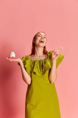 Photo for Portrait of young beautiful girl in cute dress posing with cupcake over pink background. Happy, positive vibe. Concept of youth, beauty, fashion, lifestyle, emotions, facial expression. Ad - Royalty Free Image
