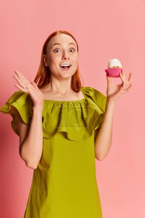 Photo for Portrait of young beautiful girl in cute dress posing with cupcake over pink background. Tasty bite. Dessert lover. Concept of youth, beauty, fashion, lifestyle, emotions, facial expression. Ad - Royalty Free Image