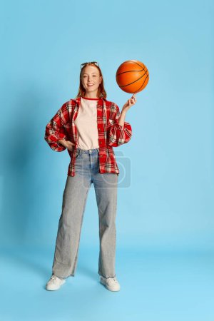 Téléchargez les photos : Portrait de jeune fille rousse en chemise à carreaux décontractée posant, tournant ballon de basket sur doigt sur fond bleu. Concept de jeunesse, beauté, mode, mode de vie, émotions, expression faciale. Publicité - en image libre de droit
