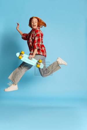 Foto de Retrato de una joven pelirroja posando en una camiseta casual a cuadros, corriendo con monopatín sobre fondo azul. Concepto de juventud, belleza, moda, estilo de vida, emociones, expresión facial. Anuncio - Imagen libre de derechos