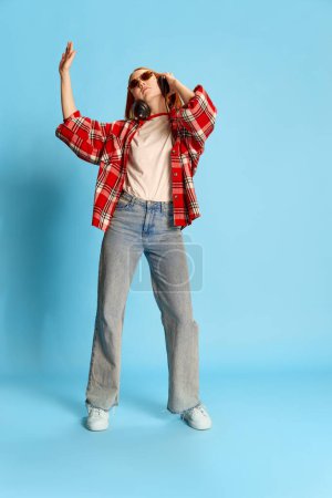 Photo for Portrait of young redhead girl in casual checkered shirt listening to music in headphones, dancing over blue background. Concept of youth, beauty, fashion, lifestyle, emotions, facial expression. Ad - Royalty Free Image