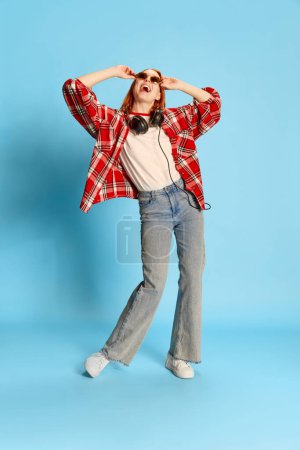 Téléchargez les photos : Portrait de jeune fille rousse en chemise à carreaux décontractée, en casque dansant sur fond bleu. Positivité. Concept de jeunesse, beauté, mode, mode de vie, émotions, expression faciale. Publicité - en image libre de droit