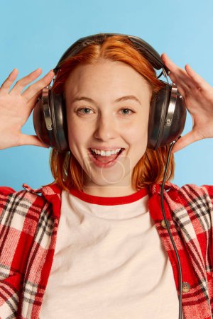 Téléchargez les photos : Portrait de jeune fille rousse en chemise à carreaux décontractée posant dans des écouteurs sur fond bleu. Heureux. Concept de jeunesse, beauté, mode, mode de vie, émotions, expression faciale. Publicité - en image libre de droit