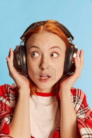 Téléchargez les photos : Portrait de jeune fille rousse en chemise à carreaux décontractée posant dans des écouteurs sur fond bleu. Un regard intéressé. Concept de jeunesse, beauté, mode, mode de vie, émotions, expression faciale. Publicité - en image libre de droit