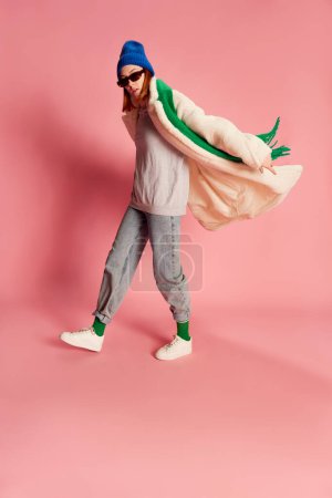 Foto de Retrato de una joven con estilo en gafas de sol, sombrero azul, bufanda verde y abrigo de piel posando sobre fondo rosa. Concepto de juventud, belleza, moda invernal, estilo de vida, emociones, expresión facial. Anuncio - Imagen libre de derechos
