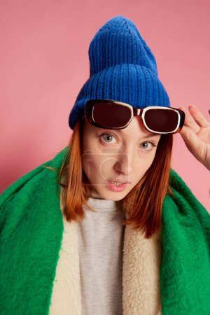 Photo for Stylish young girl in sunglasses, blue hat, green scarf and fur coat posing over pink background. Questioning look. Concept of youth, beauty, winter fashion, lifestyle, emotions, facial expression. Ad - Royalty Free Image
