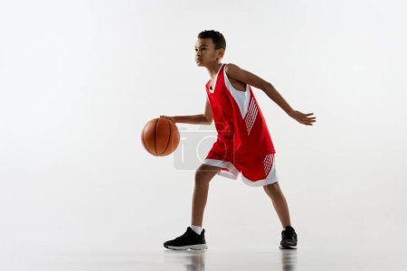 Téléchargez les photos : Portrait de garçon en uniforme rouge, jouant au basket, dribblant sur fond de studio gris. Concentration. Concept d'énergie, sport professionnel, mouvement, action, passe-temps, compétition, réalisation. - en image libre de droit