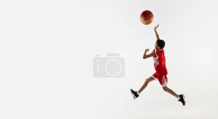 Téléchargez les photos : Portrait de garçon en uniforme rouge, jouant au basket sur fond de studio gris. Bannière. Concept d'énergie, sport professionnel, mouvement, action, passe-temps, compétition, réalisation. - en image libre de droit