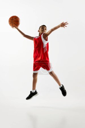 Téléchargez les photos : Portrait d'un garçon en uniforme rouge s'entraînant, jouant au basket, lançant une balle dans un saut par-dessus un fond de studio gris. L'objectif. Concept d'énergie, sport professionnel, mouvement, passe-temps, compétition, réalisation. - en image libre de droit