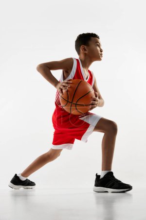 Foto de Retrato de niño en uniforme rojo entrenando, jugando baloncesto sobre fondo gris estudio. Preparación. Concepto de energía, deporte profesional, movimiento, acción, hobby, competición, logro. - Imagen libre de derechos