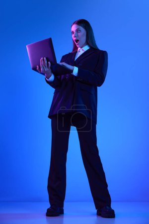 Foto de Mujer en traje oficial elegante que trabaja en el ordenador portátil sobre fondo azul en luz de neón. Empleado sorprendido. Concepto de empresa, tecnologías modernas, ocupación, desarrollo profesional - Imagen libre de derechos