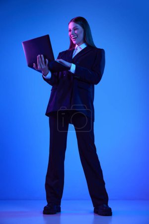 Téléchargez les photos : Femme en costume officiel élégant travaillant sur ordinateur portable sur fond bleu dans la lumière au néon. Une coopération réussie. Concept d'entreprise, technologies modernes, profession, évolution de carrière - en image libre de droit