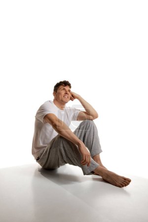 Photo for Relaxed look. Mature smiling man sitting on floor in T-shirt and pants over white studio background. Homewear. Comfort. Concept of mens health and beauty, body and skin care, fitness. Body art - Royalty Free Image