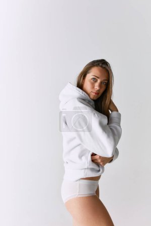 Foto de Portrait of beautiful woman posing in white hoodie over grey studio background. Feeling comfortable. Self-care. Concept of body and skin care, fitness, natural beauty, health, wellness. - Imagen libre de derechos