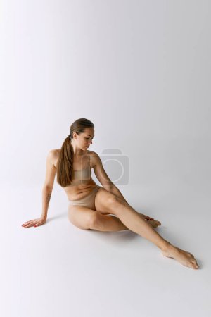 Téléchargez les photos : Full-length portrait of beautiful woman posing in beige underwear over grey studio background. Slender body. Concept of body and skin care, fitness, natural beauty, health, wellness. - en image libre de droit