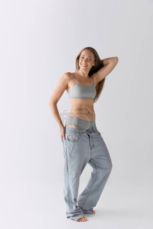 Photo for Full-length portrait of slim woman posing in top and jeans over grey studio background. Positive, smiling mood. Concept of body and skin care, fitness, natural beauty, health, wellness. - Royalty Free Image