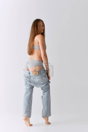 Téléchargez les photos : Full-length portrait of slim woman posing in top and jeans over grey studio background. Sself-love and acceptance. Concept of body and skin care, fitness, natural beauty, health, wellness. - en image libre de droit
