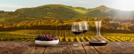Téléchargez les photos : Decanter and two glasses with delicious red wine and grape appetizers standing on wooden table over mountain background with field. Countryside winemaking. Traditional taste of natural wine - en image libre de droit