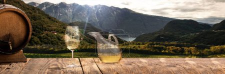 Foto de Decanter, glass with white wine and wooden barrel standing on wooden table over beautiful mountain and field landscape. Winemaking industry, degustation. Traditional taste of natural alcohol drink - Imagen libre de derechos