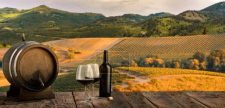 Téléchargez les photos : Decanter, two glasses with red wine and wooden barrel standing on wooden table over beautiful mountain and field landscape in summertime. Countryside winemaking. Traditional taste of natural wine - en image libre de droit