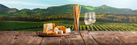 Foto de Two glasses of delicious white wine with cheese board appetizers standing on wooden table. Beautiful field view in summer. Romantic date in countryside. Traditional taste, wine making concept - Imagen libre de derechos