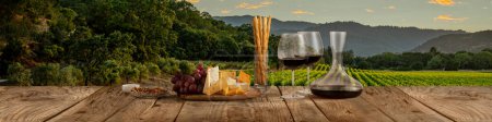 Téléchargez les photos : Decanter and two glasses with delicious red wine, grape and cheese appetizers standing on wooden table over beautiful nature landscape of fields and forest. Traditional taste, wine making concept - en image libre de droit