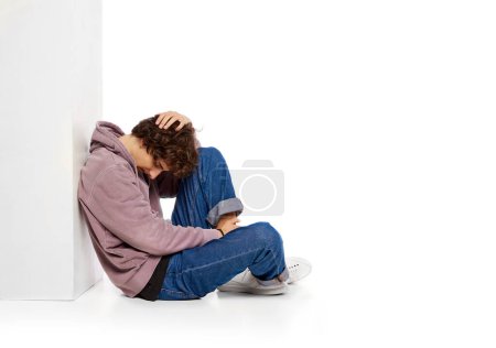 Foto de Portrait of young boy sitting on floor in depression and sadness over white background. Problems of youth. Concept of psychology, inner world, mental health, emotions, feelings - Imagen libre de derechos