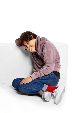 Téléchargez les photos : Portrait of young boy sitting on floor in despair and sadness over white background. Suffering from depression and breakdown. Concept of psychology, inner world, mental health, emotions, feelings - en image libre de droit