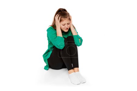 Foto de Portrait of depressed teen girl sitting on floor and covering ears with hands over white background. Breakdown, apation. Concept of psychology, inner world, mental health, emotions, feelings - Imagen libre de derechos