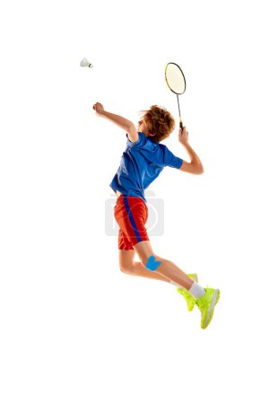 Téléchargez les photos : Portrait of teen boy in uniform playing badminton, serving shuttlecock with racket in a jump isolated over white background. Concept of sportive lifestyle, motion, action, competition - en image libre de droit