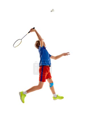 Téléchargez les photos : Portrait of teen boy in uniform, badminton player hitting shuttlecock with racket in a jump, training isolated over white background. Concept of sportive lifestyle, motion, action, competition - en image libre de droit