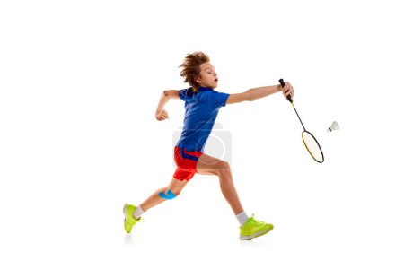 Téléchargez les photos : Portrait of teen boy in uniform playing badminton, hitting shuttlecock with racket in a run isolated over white background. Concept of sportive lifestyle, motion, action, competition - en image libre de droit