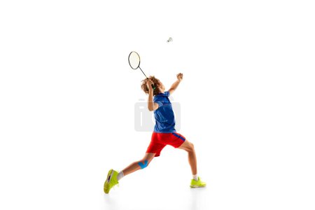 Téléchargez les photos : Portrait of teen boy in uniform in motion, playing badminton, serving shuttlecock with racket isolated over white background. Concept of sportive lifestyle, motion, action, competition, hobby - en image libre de droit