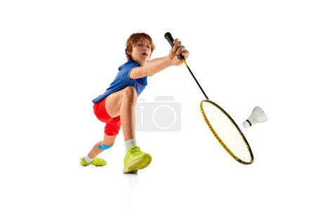 Téléchargez les photos : Portrait of concentrated teen boy in uniform playing badminton, serving shuttlecock with racket isolated over white background. Concept of sportive lifestyle, motion, action, competition - en image libre de droit