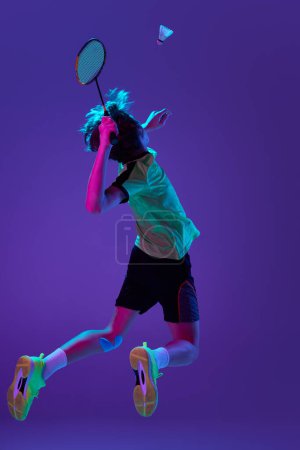 Photo for Back view. Portrait of teen boy in uniform playing badminton, hitting shuttlecock in a jump over blue purple background in neon ligth. Concept of sportive lifestyle, motion, action, competition - Royalty Free Image